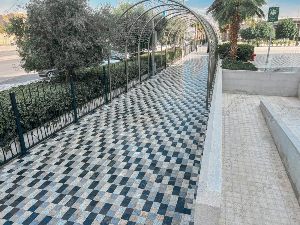 An outdoor pathway covered with a metal dome and tiled with a checkerboard pattern. The pathway is surrounded by olive and palm trees, with several shrubs on the opposite side. A road and more greenery are visible in the background.