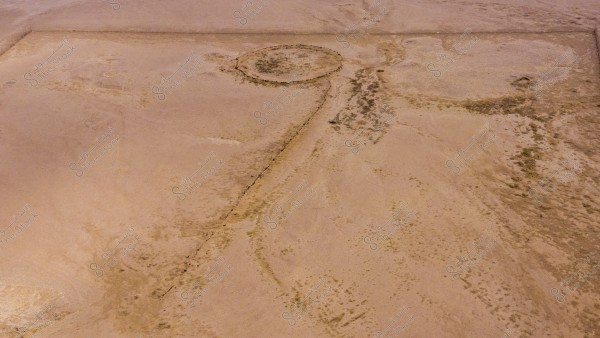 An aerial view of a desert landscape featuring a prominent circular formation on the ground, from which a straight line extends. The terrain is sandy brown with sparse dry vegetation scattered randomly.