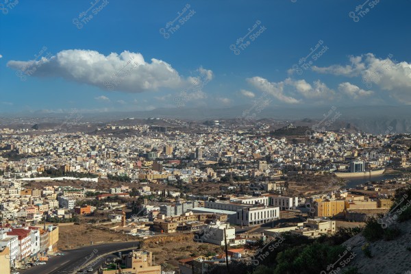 منظر طبيعي لمدينة أبها في المملكة العربية السعودية, حيث يظهر عدد كبير من المباني والبيوت المنتشرة على سفوح الجبال. السماء صافية وزرقاء مع وجود بعض السحب البيضاء الخفيفة.