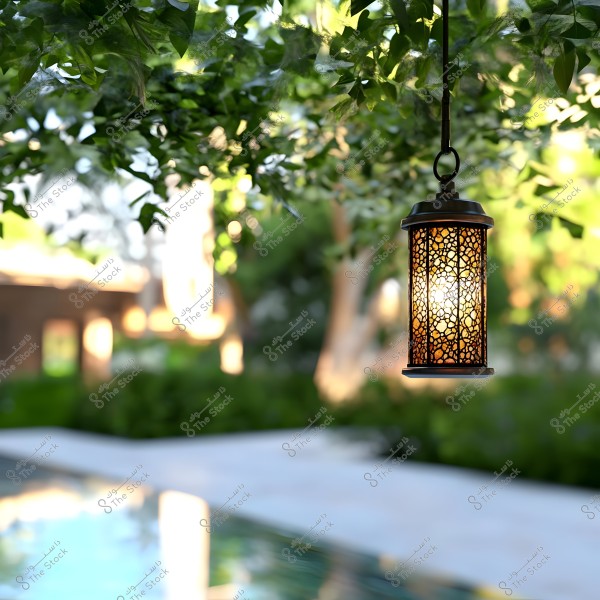 A decorative metal lantern hanging from a tree outdoors. The lantern glows with a warm light, surrounded by green foliage, with a blurred background of a garden and a pool reflecting the light.