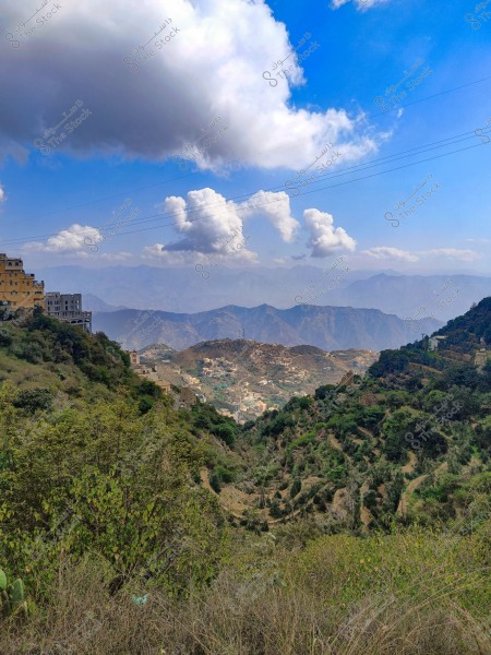 منظر طبيعي لقرية جبلية تحيط بها النباتات الخضراء الكثيفة. تظهر في الخلفية سلسلة من الجبال مع سماء زرقاء صافية تتخللها سحب بيضاء كبيرة. على الجانب الأيسر, يمكن رؤية مبانٍ تطل على السفوح الخضراء.