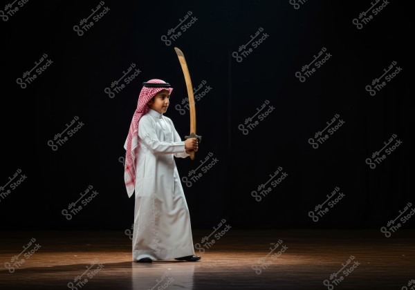 A photo of a young child wearing traditional Saudi attire, a white thobe, a red-checkered ghutra, and a black agal, standing on a stage holding a wooden sword. The background is black, with a spotlight beautifully illuminating him.