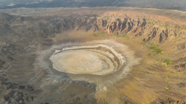 An aerial view of a volcanic crater in a mountainous desert region, featuring a large circular shape surrounded by wavy lines that appear to be mineral deposits. The area is surrounded by hills and mountains in brown and gray hues, with patches of green vegetation scattered across parts of the landscape. The horizon stretches out in the background, adding depth to the natural scene.