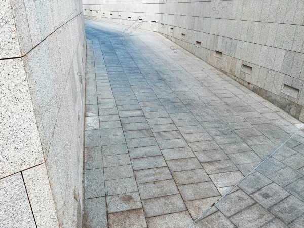 The image shows a sloped pathway covered with stone tiles running between concrete walls. The walls are close together, forming a narrow, curved path that appears clean and modern in design.