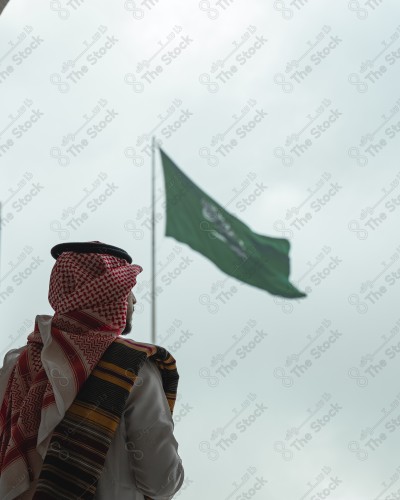 A Saudi man in traditional Saudi dress stands tall in front of the Saudi flag above a building in Rijal Almaa village during the day in the Asir region. A Saudi man celebrates the National Day, the Saudi National Day.