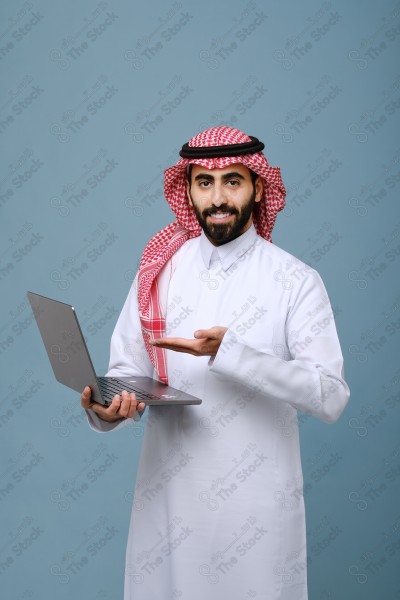 بورتريه شاب سعودي يرتدي الزي السعودي يأشر إلى جهاز المحمول وتظهر على ملامحة السعادة ، يقف على خلفية سماوية