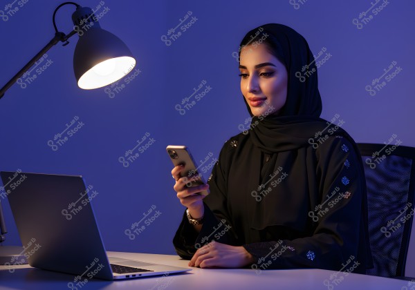 A portrait of a woman wearing a black abaya and hijab, sitting at a desk and using a smartphone. In front of her is a laptop illuminated by a desk lamp. The blue background creates a calm atmosphere.