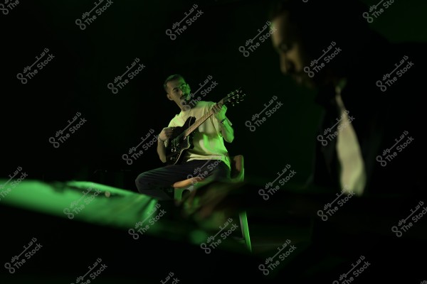 شخص يعزف على الغيتار في إضاءة خضراء خافتة مع شخص آخر في الخلفية غير واضح.