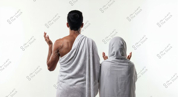 صورة تظهر شخصين من الخلف بملابس الإحرام, الشخص الأول يرتدي إزار أبيض مكشوف الكتف الأيمن, والثاني يرتدي حجاب وعباءة بيضاء, يرفعان أيديهما بالدعاء. خلفية الصورة بيضاء ومسطحة.