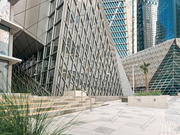 An image of a modern complex with distinctive architectural design, featuring slanted glass walls and metallic elements. The buildings are surrounded by a wide, clean paved area with some green plants and nearby palm trees. The glass facade reflects the neighboring skyscrapers on a sunny day.
