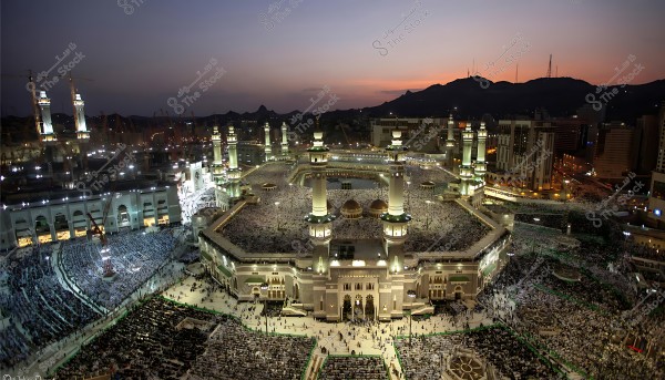 منظر جوي للمسجد الحرام في مكة المكرمة أثناء الغروب, يحيط به المصلون في الساحات الخارجية.