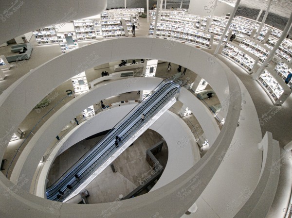 An image showing an interior view of a modern library with a circular architectural design. The library features multiple spiral floors with well-lit bookshelves. There is an escalator in the center that moves visitors between the floors. The design is spacious and contemporary, with bright lighting and open space.