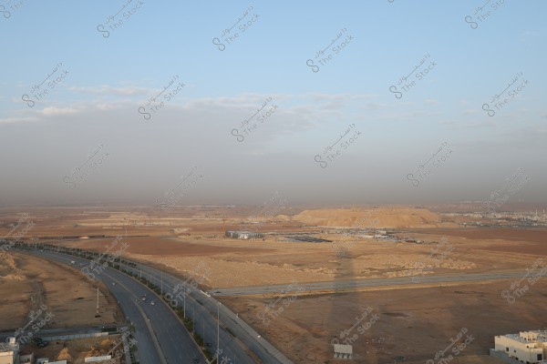 Image of a desert landscape featuring a wide road stretching through open, arid areas with sand dunes visible in the distance under a partly clear blue sky. Small buildings and industrial areas can be seen in the distance, with some wispy clouds in the sky.