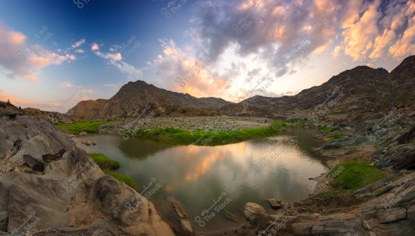منظر طبيعي لبحيرة صغيرة محاطة بصخور وجبال تحت سماء غائمة جزئياً عند الغروب. تتنوع الألوان بين الأزرق والبرتقالي, وتنبت الأعشاب الخضراء على ضفاف البحيرة مما يضفي حيوية على المشهد.