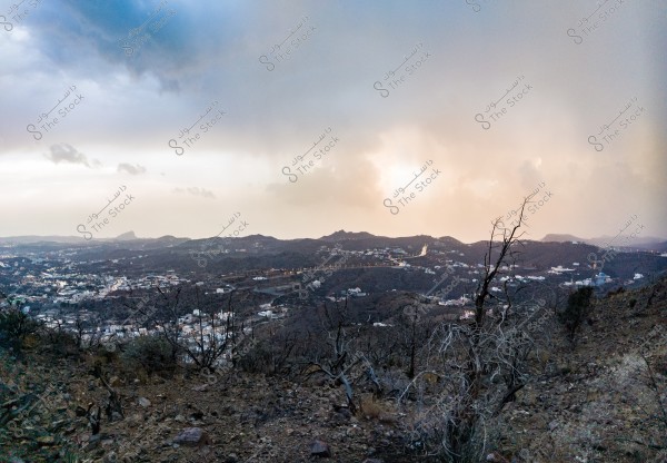 Scenic view of a mountain in Saudi Arabia, showcasing a vast landscape with a range of mountains and scattered hills under a partially cloudy sky. White buildings are interspersed across the rugged terrain, with dry trees visible in the foreground.