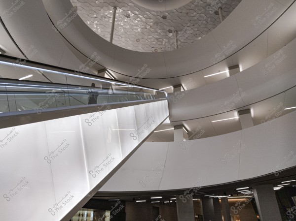 An image of a modern building interior, featuring an escalator inclined upwards with glass and metal walls reflecting the ambient lighting. The escalator is surrounded by circular walls and a structure with geometric patterns on the ceiling. The lighting and modern design create a futuristic atmosphere.