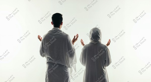 The image shows two individuals standing from behind, wearing white Ihram garments, indicating they are preparing for Hajj or Umrah rituals. The person on the left appears to be a man with short hair, while the person on the right appears to be a woman wearing a headscarf that covers her head. Both have their hands slightly raised in a prayer position. The simple white background adds a spiritual and serene atmosphere to the scene.