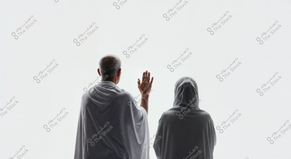 يظهر في الصورة شخصان من الخلف, يرتديان ملابس الإحرام البيضاء. الشخص الأول على اليسار يبدو أكبر سناً ويرفع يده اليمنى في وضع الدعاء, بينما يقف الشخص الثاني على اليمين مرتديًا حجابًا يغطي الرأس. الخلفية بيضاء بسيطة تضفي إحساسا بالسكون والتأمل.
