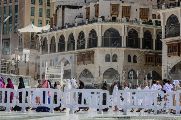 A group of people dressed in white Ihram garments and colorful Islamic abayas walking in the Haram area in Mecca, Saudi Arabia. Behind them are buildings with traditional Islamic architectural designs featuring arched windows. The weather is sunny, and there appears to be a refreshing breeze.