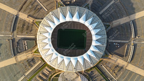 Aerial view of a large stadium in Saudi Arabia, showcasing the distinctive circular roof design resembling white sun rays. The stadium is surrounded by neatly organized parking lots and roads. The green field inside the stadium is visible, indicating the playing area.