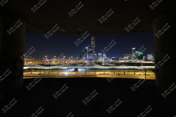 Night view of a modern city with illuminated skyscrapers in the background. The foreground features a highway with light traffic and several elevated bridges. The background buildings include skyscrapers with contemporary architecture and striking lighting details, suggesting a modern and thriving urban area.