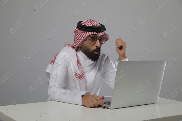 Image of a man sitting in front of a laptop on a white table. The man is wearing traditional Saudi attire, including a white thobe and a red-and-white checkered ghutra with a black agal. He appears surprised or focused as he looks at the screen.