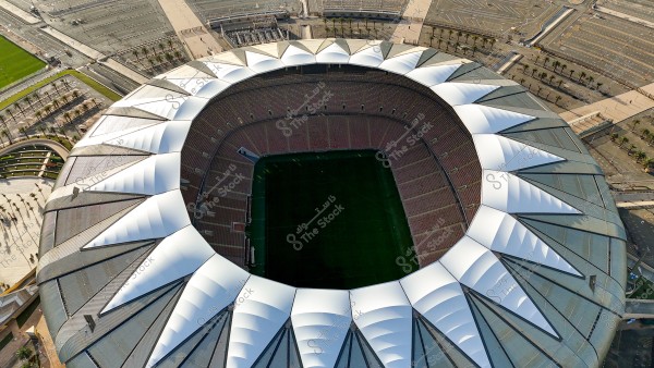 Aerial view of a large sports stadium with distinctive architectural design, featuring a circular top covered with white structures resembling sails. The vast seating areas descend gradually towards the green field in the center. The stadium is surrounded by parking lots, green areas, and palm trees.
