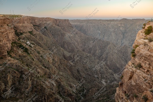 An expressive image of a deep valley with steep mountain sides in a desert area. The mountain rocks appear dry and rugged, with some small shrubs scattered on the slopes. The sky is a pale blue towards the horizon, with faint lighting showing remnants of sunset.