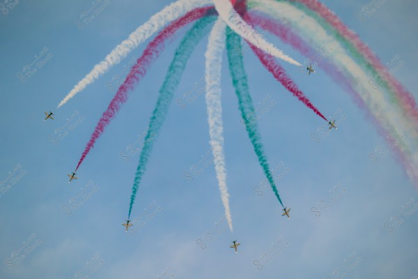 Aircraft performing an aerial display in the sky, creating smoke trails in white, red, and green colors. The aircraft are arranged in an artistic formation against a blue sky background.