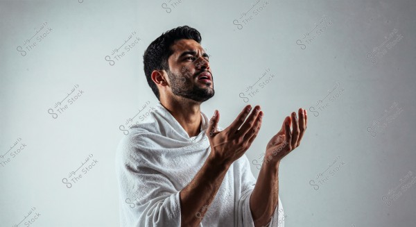 A portrait of a man appearing to be in a prayer or supplication position, wearing a white Ihram garment. His face is turned upwards with his hands raised. The background is light gray.