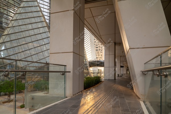 The image shows a modern architectural scene with angular design, featuring pathways adorned with glass and metal. Large triangular glass structures composed of multiple glass panels are visible in the background. A pedestrian walkway with glass barriers and metal railings runs along the left side, while sunlight enters at an angle, adding depth and warm lighting to the area.