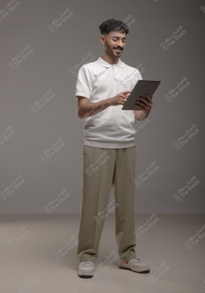 A man stands in a studio, wearing a white polo shirt and beige pants, holding a tablet and appearing to smile. The background is gray.