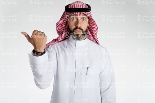 بورتريه رجل سعودي يقف على خلفية بيضاء، يرتدي الثوب، ينظر الى الكاميرا ويقوم بعمل حركات بيديه، تعبير اندهاش وتفاجؤ