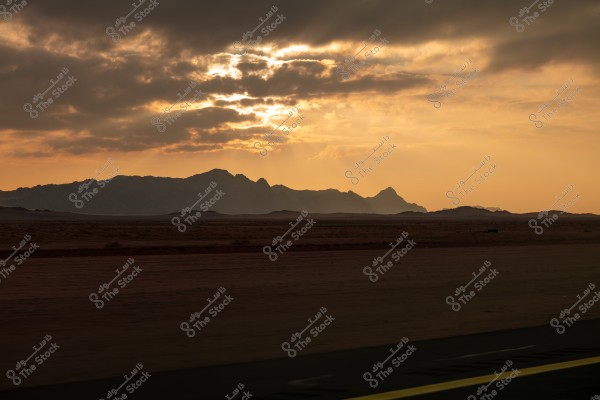 Image of a sunset behind a mountain range in a desert area. The sky is filled with dark clouds with warm sunlight filtering through, casting golden and orange hues over the landscape. The ground is dry and flat with a visible road in the foreground.