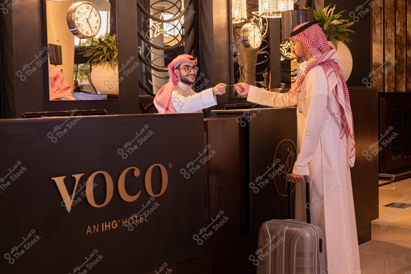 مشهد داخلي لمكتب استقبال في فندق. يظهر في الصورة موظف استقبال يقف خلف المكتب يرتدي ثوباً وغترة وعقال, ويتبادل التحية مع نزيل يقف على الجانب الآخر يرتدي أيضاً الزي التقليدي ويظهر وهو يسلم بطاقة. توجد ساعة حائط ونبات زينة على المكتب, مع شعار فندق \"فووكو\" واضح في المقدمة.