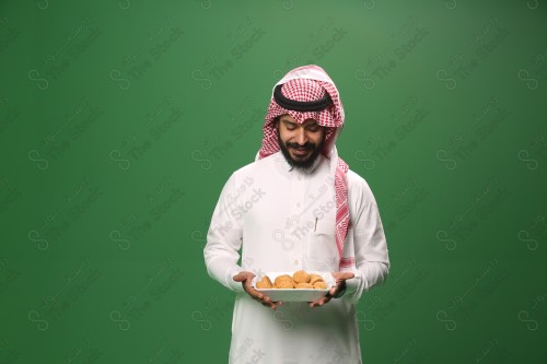 شاب سعودي على خلفية خضراء يرتدي الثوب و الشماغ، يقوم بعمل حركات بيده،يمسك بصينية الكعك و يقدمه، احتفالات العيد