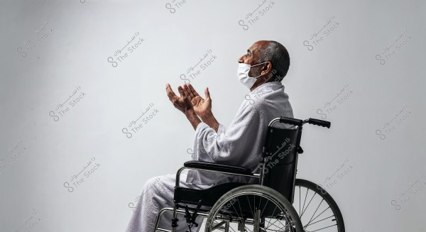 An image of an elderly man sitting in a wheelchair, raising his hands in prayer. He is wearing a white robe and a surgical mask. The background is white, highlighting the man\'s side profile and calm demeanor.