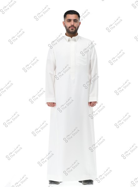 An image of a man standing in a studio wearing a long white garment known as a thobe, a traditional attire popular in Gulf countries. The man is wearing brown shoes, and the background is completely white.