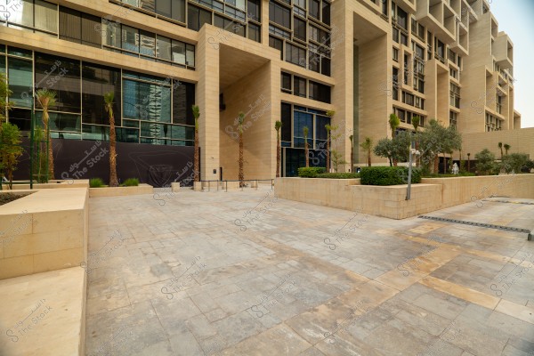 An image showing a modern stone plaza surrounded by tall buildings with a contemporary architectural design of stone and glass. Palm trees and bushes are neatly arranged on the sides. The plaza is clean and spacious.