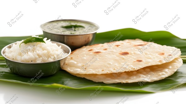 Image of a selection of Indian food items resting on a green banana leaf. The image includes a small metal bowl containing white rice garnished with curry leaves, and another bowl with green sauce. Next to them, there are thin flatbread pieces.