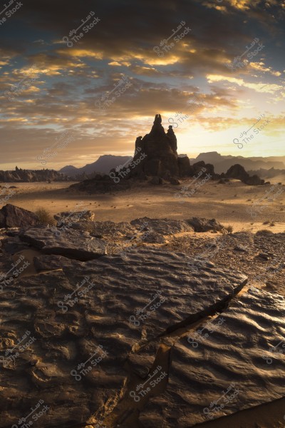 A desert landscape featuring large rock formations in the foreground with a tall mountain in the background. The sky is filled with clouds with gradients of grey and yellow, suggesting a sunset. The ground is covered with sand and scattered rocks.