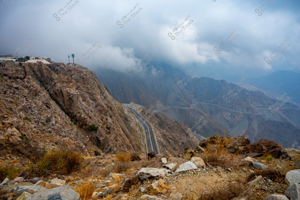 A natural landscape of towering mountains in Saudi Arabia with a paved road winding along the mountains. The barren rocks are in shades of brown and gray, while thick clouds cover the mountain peaks in the sky. A few small buildings and palm trees can be seen at the top side of the image.
