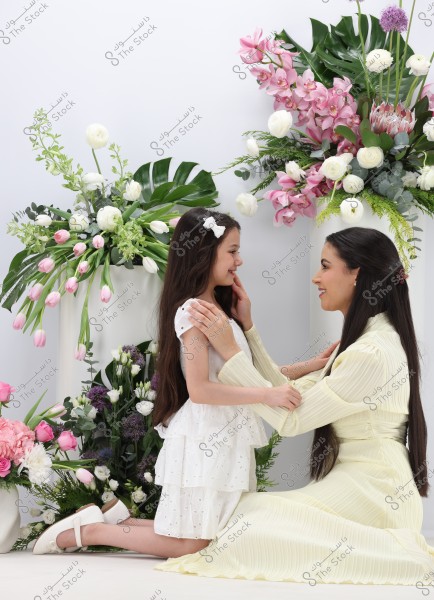 An image of a woman in a light-colored long dress kneeling in front of a young girl in a white dress. The girl is smiling and the woman is gently touching her face. Behind them are floral arrangements of various flowers in white, pink, and green.