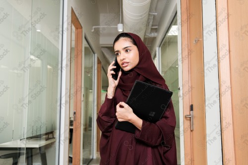 Saudi woman wearing an abaya stands in the meeting room in front of the glass front, holding a tablet ، during the day