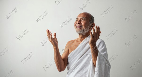 An elderly man wearing a white Ihram, a garment associated with Islamic culture, set against a simple white background. The man appears smiling and has his hands raised in a gesture of prayer or meditation.