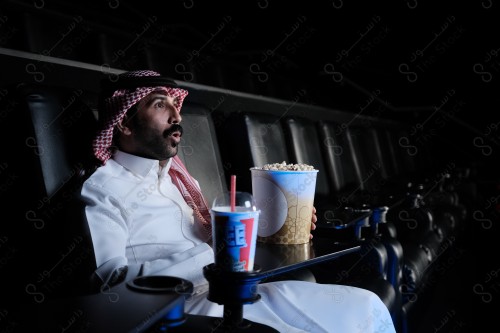 شاب سعودي يرتدي الزي السعودي التقليدي يشاهد أحد الأفلام المعروضة في صالة السينما بينما يبدو مندهشًا ويظهر أمامه الفشار وأحد المشروبات على الطاولة