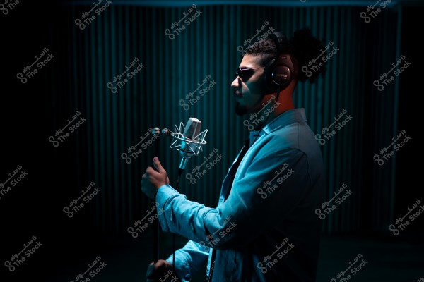 رجل يرتدي سماعات رأس ونظارات شمسية يغني في ميكروفون في استوديو مظلم.