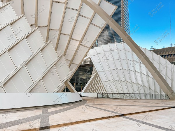 An image of a modern architectural structure composed of white panels arranged at an angle, with metallic intersections. The building is surrounded by a light-tiled floor, and a clear blue sky is in the background. In the far corner, a tall building with glass panels is visible.