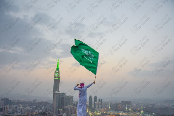 A snapshot of a Saudi man in traditional Saudi dress holding the Saudi flag over a mountain in the Makkah Al-Mukarramah region, the sky, the royal clock tower building in the Grand Mosque, buildings and landmarks, the Grand Mosque of Mecca.