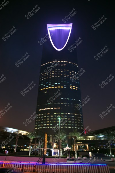 A nighttime view of the Kingdom Tower in Riyadh, Saudi Arabia. The tower is known for its unique architectural design, featuring a purple-lit arch at its top. Palm trees and city lights are visible in the foreground.